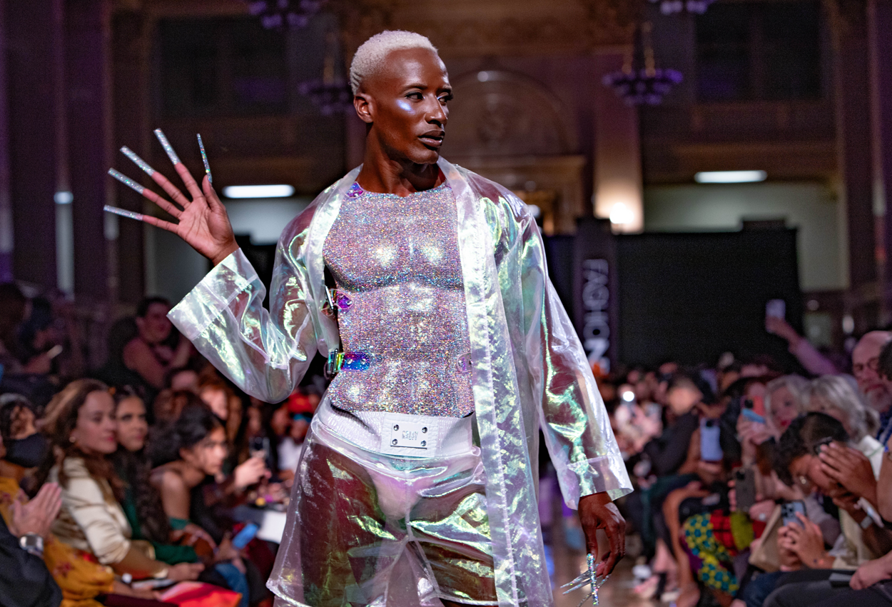 A model with short platinum hair walks a runway wearing a shimmering, iridescent sheer coat and shorts over a sparkling top, striking a dramatic pose with long decorated nails. An audience watches attentively.