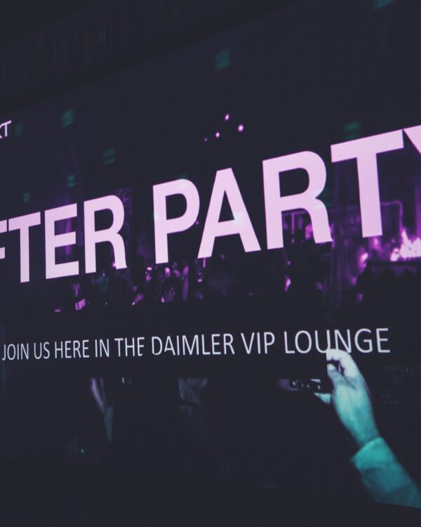 Large screen with purple text reading "AFTER PARTY" and smaller text below saying "JOIN US HERE IN THE DAIMLER VIP LOUNGE," with a crowd and someone holding a phone in the foreground. Large screen with purple text reading "AFTER PARTY" and smaller text below saying "JOIN US HERE IN THE DAIMLER VIP LOUNGE," with a crowd and someone holding a phone in the foreground.