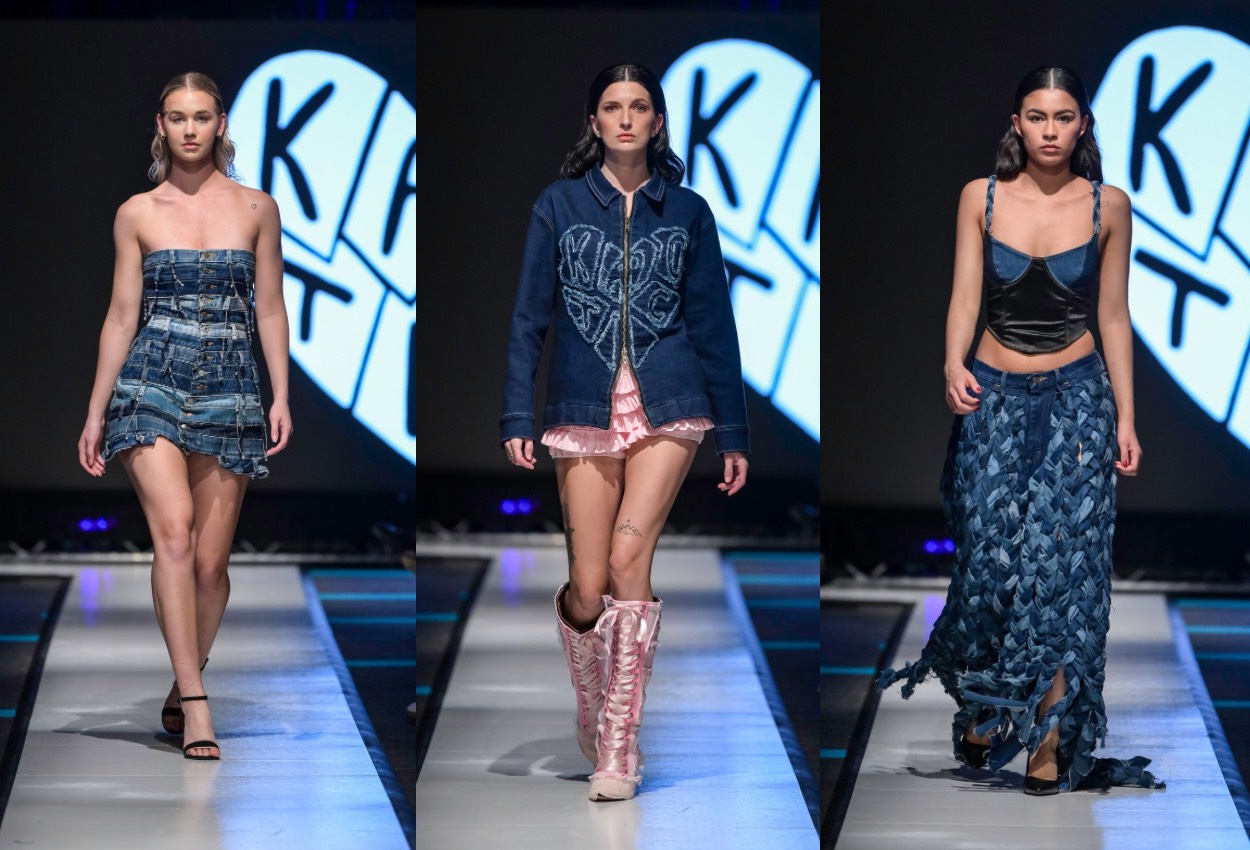 Three models walk a runway wearing blue denim-inspired outfits: a strapless mini dress, a heart-embroidered jacket with pink shorts and boots, and a woven skirt with a black top. The background features a bold black-and-white logo.