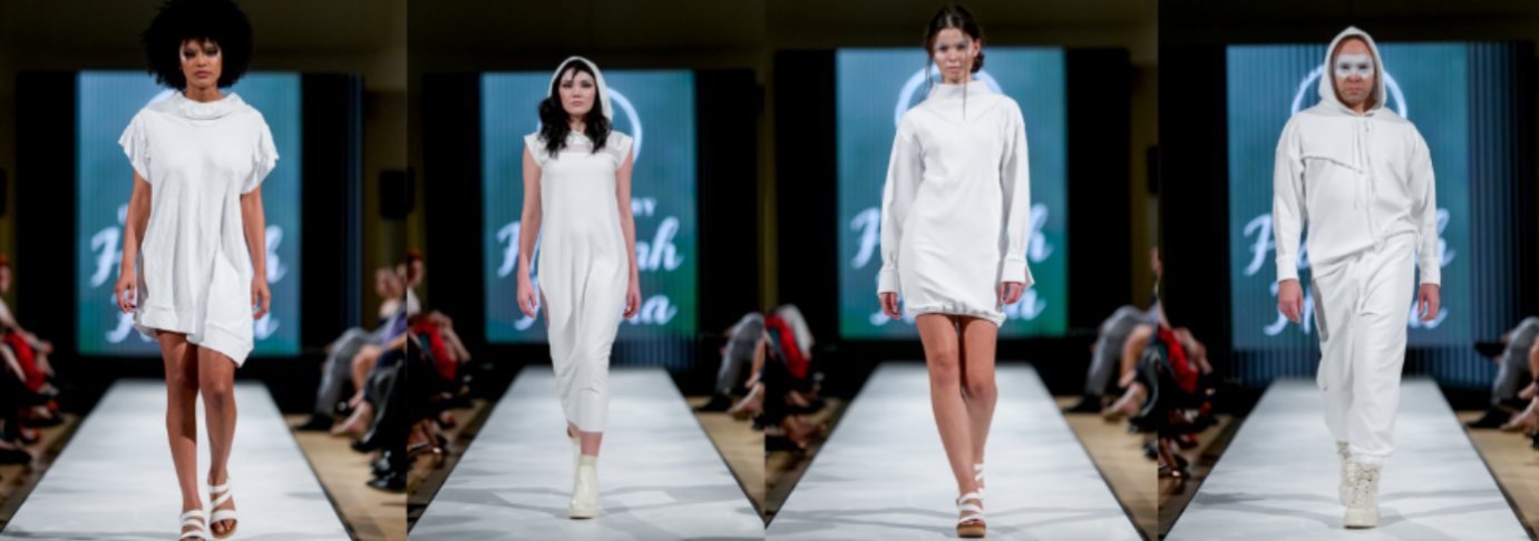 Four models walk down a runway wearing minimalist, all-white outfits, including dresses and a hooded ensemble, as an audience watches from both sides. A screen with blurred text is visible in the background.