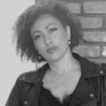 Black and white photo of a woman with curly hair, wearing a dark jacket and earrings, standing in front of a brick wall, looking at the camera with a neutral expression.