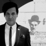 A man in a suit stands under an umbrella next to a wall with graffiti of a face wearing a hat and the words “Respect yourself.” The image is black and white.