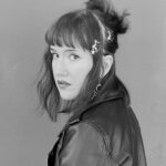 Black and white photo of a person with shoulder-length hair, wearing a leather jacket. Their hair is styled with star-shaped hair clips, and they are looking over their shoulder toward the camera, against a plain background. Black and white photo of a person with shoulder-length hair, wearing a leather jacket. Their hair is styled with star-shaped hair clips, and they are looking over their shoulder toward the camera, against a plain background.