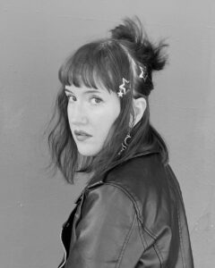 Black and white photo of a person with shoulder-length hair, wearing a leather jacket. Their hair is styled with star-shaped hair clips, and they are looking over their shoulder toward the camera, against a plain background.
