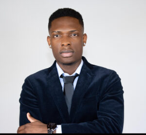 A man in a dark blue suit, white shirt, and black tie stands with his arms crossed, looking serious. He has short hair, earrings, and wears a wristwatch, posed against a plain light background.
