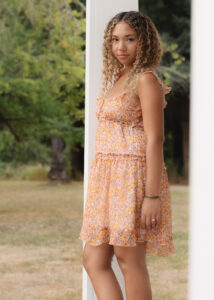 A young woman with curly hair wears a sleeveless, floral dress and stands outdoors by a white post, looking at the camera, with green trees and grass in the background.