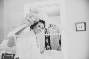 A smiling woman in a white robe holds a clothes hanger toward the camera in a bright room, with clothing visible in the background.