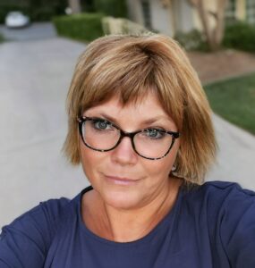 A woman with short blonde hair and glasses wearing a navy blue top stands outside on a driveway with greenery in the background, looking at the camera with a neutral expression.