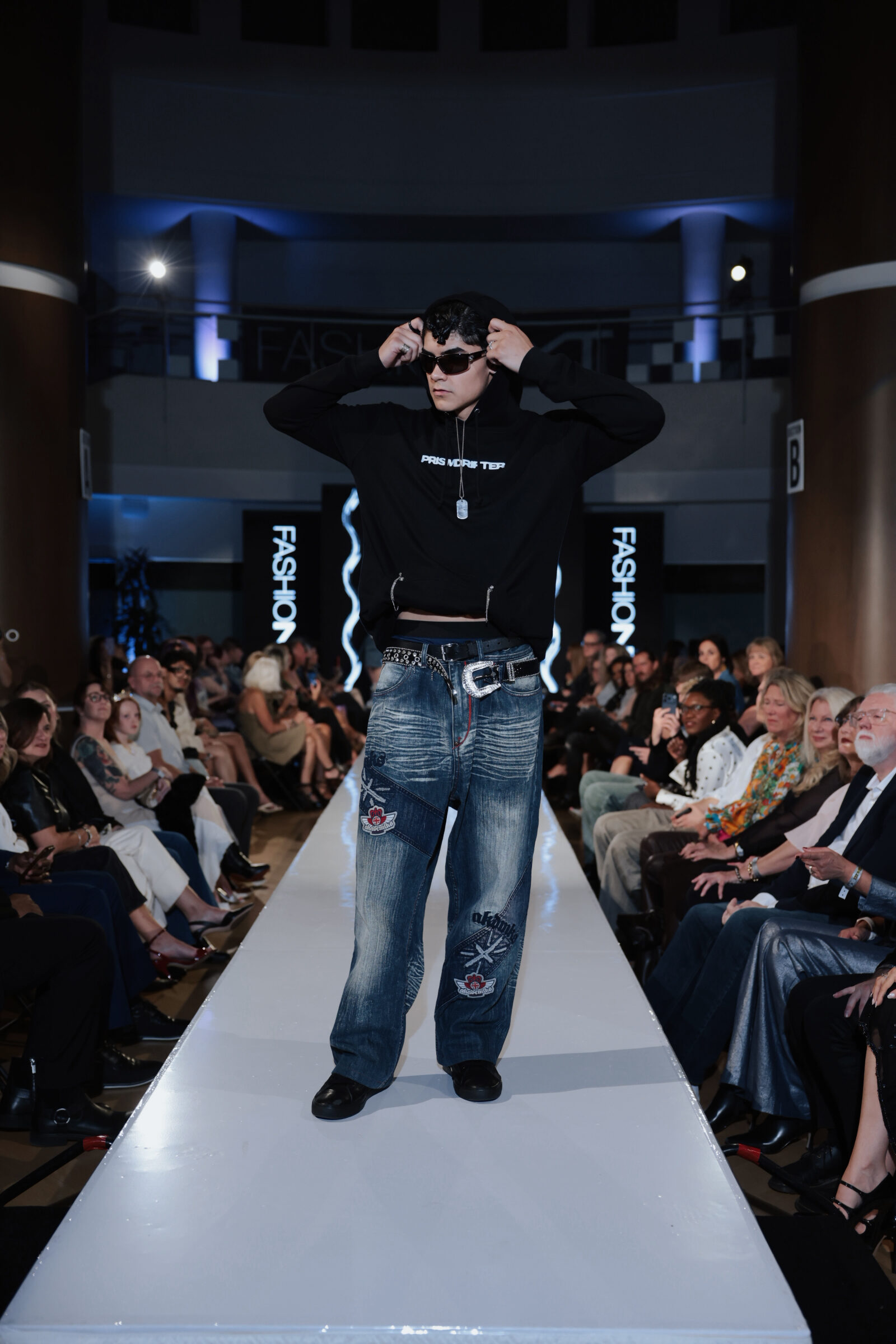 A model in a black hoodie and wide-leg jeans walks down a fashion runway in front of an audience.