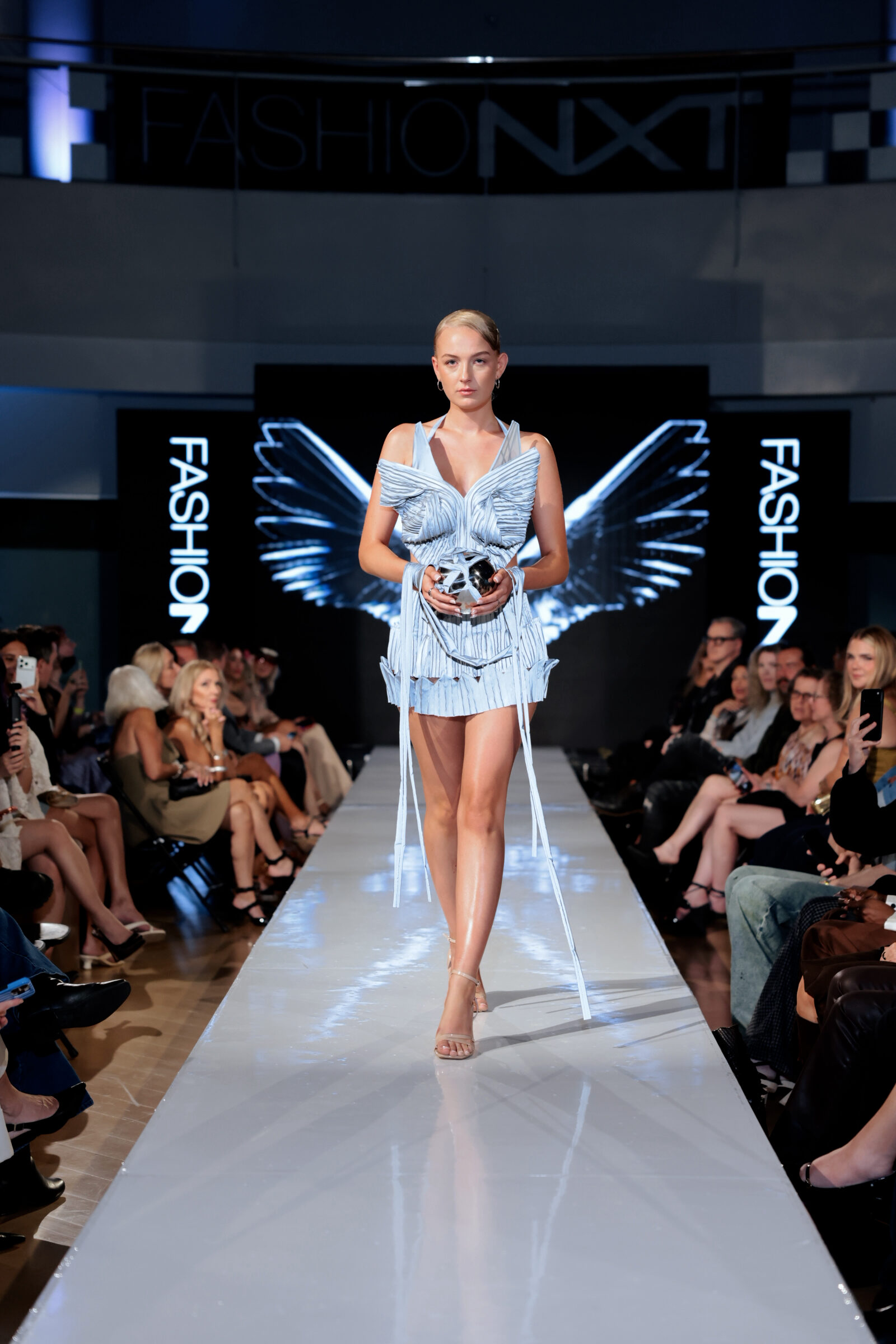 A model walks down a runway wearing a pale blue structured mini dress with vertical pleats and long ribbons, holding a silver orb.