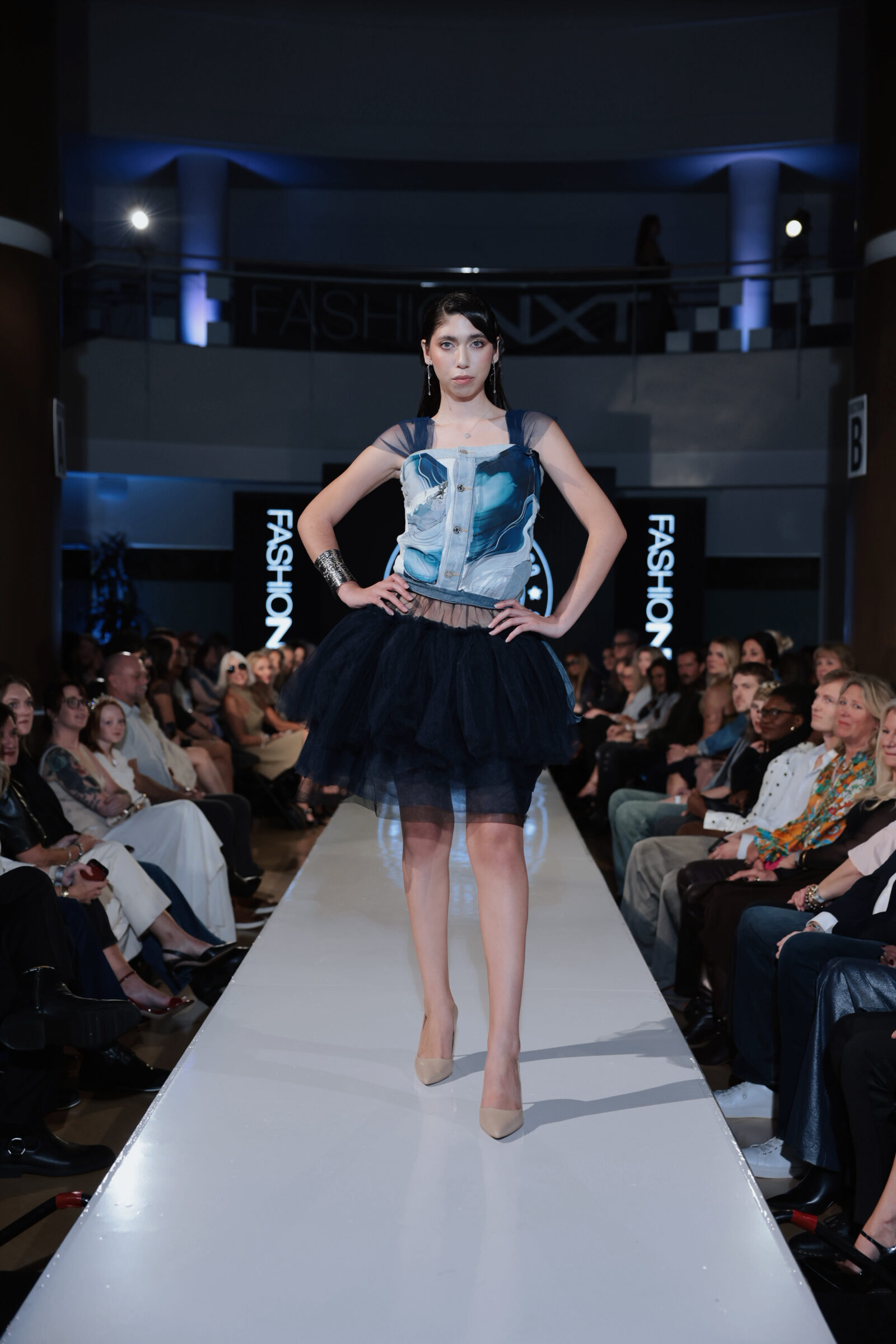A model walks a runway wearing a sleeveless top with a blue marbled pattern and a black layered tulle skirt.