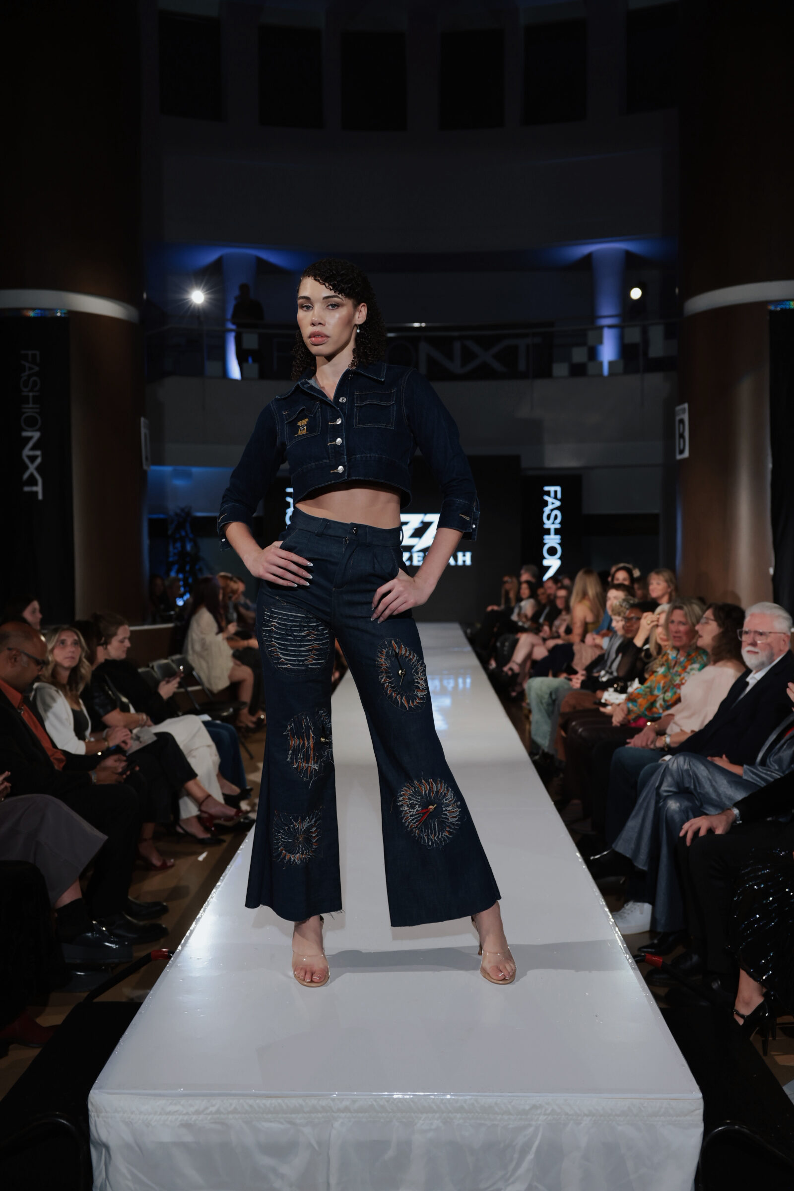 A model walks a runway in a dark denim cropped jacket and matching flared jeans embroidered with sunburst patterns.